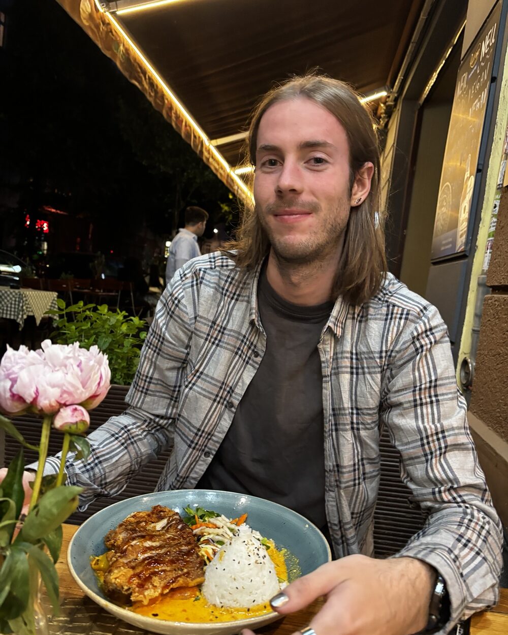Niko in a plaid shirt at an outdoor restaurant table at night, a bowl of food in front of him and pink peonies beside it
