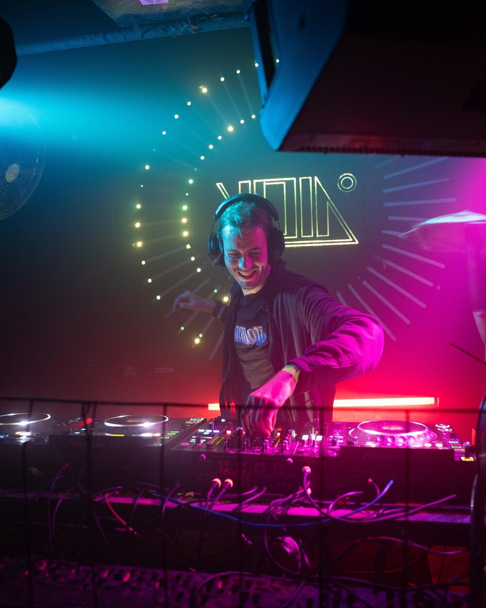 Niko DJing behind the decks in a club, lit magenta and teal, a glowing VOID backdrop behind him