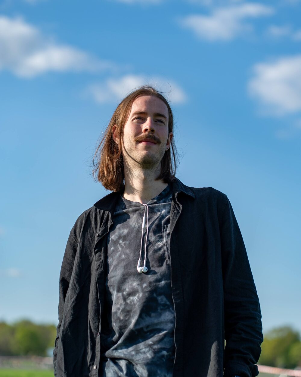 Niko outdoors on a sunny day against a bright blue sky, in a dark jacket over a tie-dye hoodie