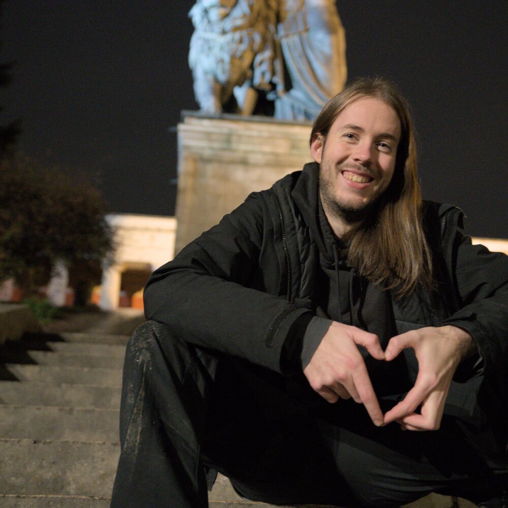 Niko sitting on stone steps at night in front of a floodlit statue, smiling and making a heart with his hands