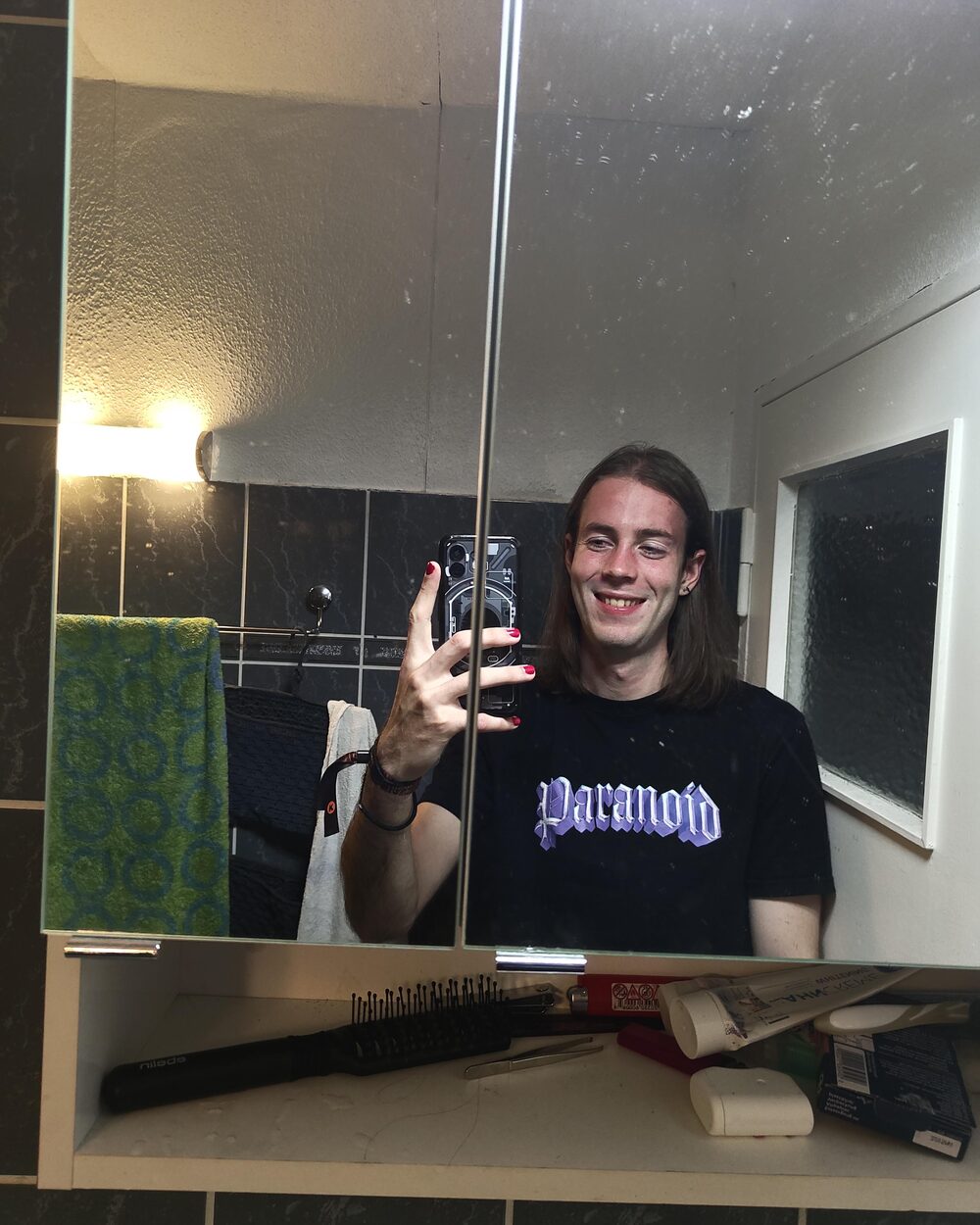 Bathroom mirror selfie, Niko in a black Paranoid t-shirt with painted nails, waving at the mirror